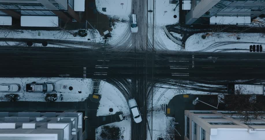 Slow descent towards vehicles driving through a snowy intersection between office buildings