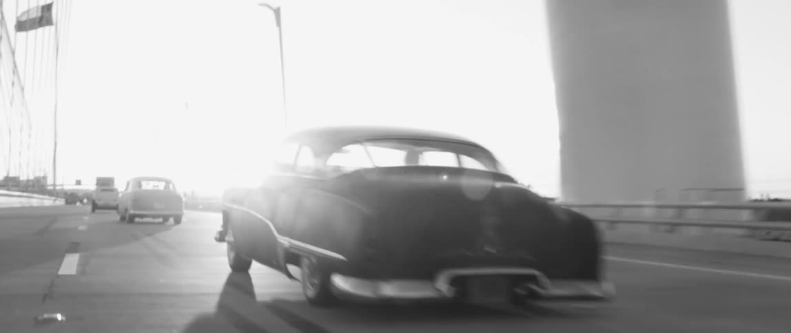 Black and white footage of vintage vehicles driving over a bridge at sunset