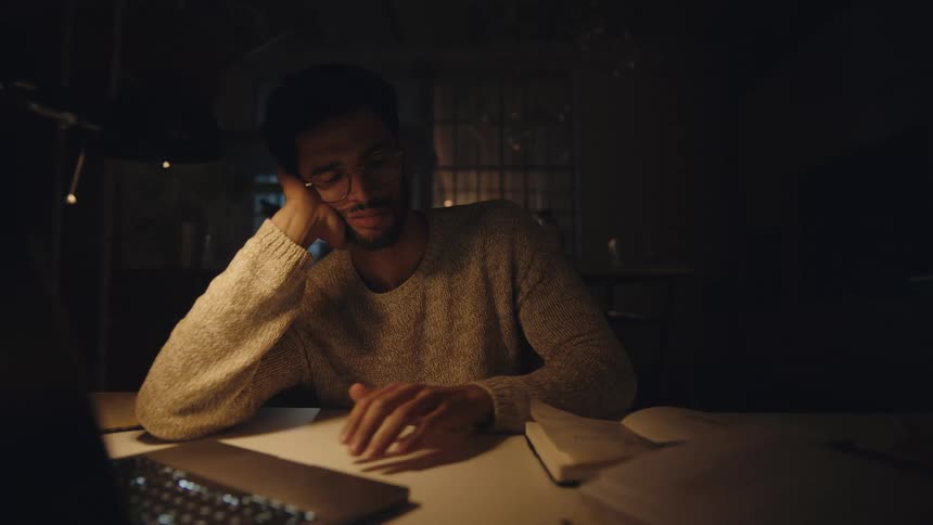 A man taps the keyboard of a laptop before looking through a notebook while sitting at a table in a dark room