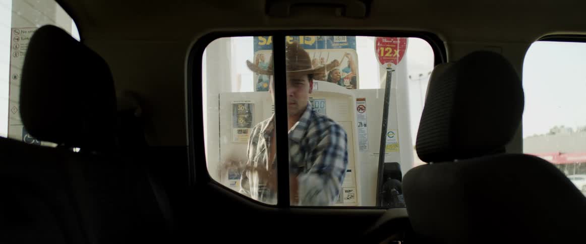 Man puts on a cowboy hat as he stands next to his vehicle and prepares to put gas in it from a gas pump