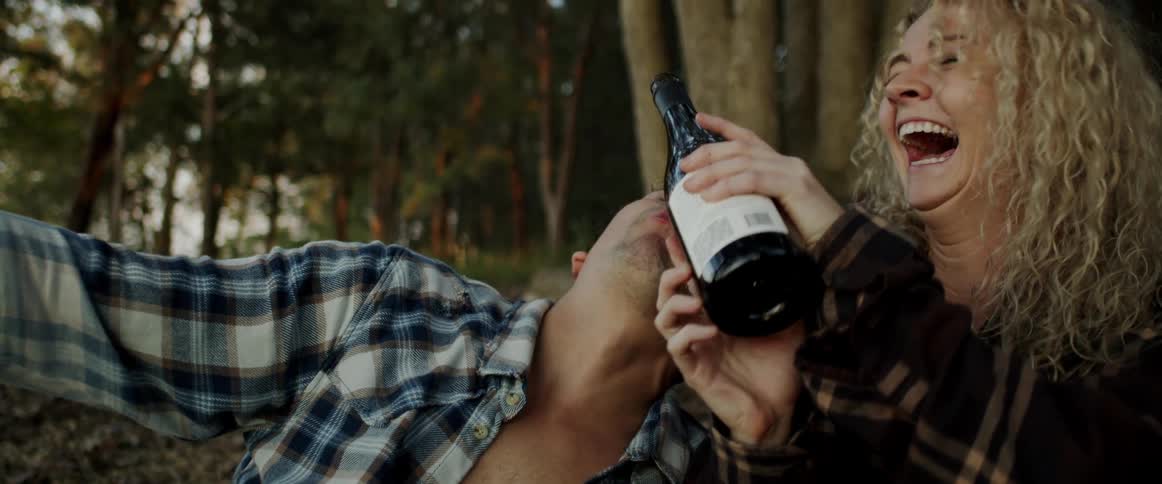 Woman pours from wine bottle into mans mouth then they laugh together as they sit on ground near trees