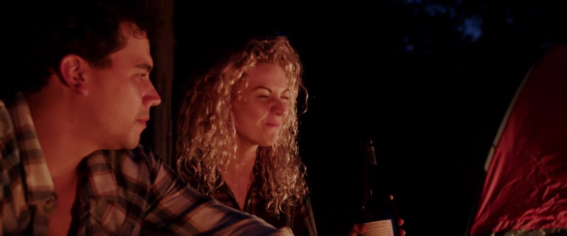 Woman drinks from a bottle of wine and laughs then hands the bottle to a man while they are camping at night in a forest