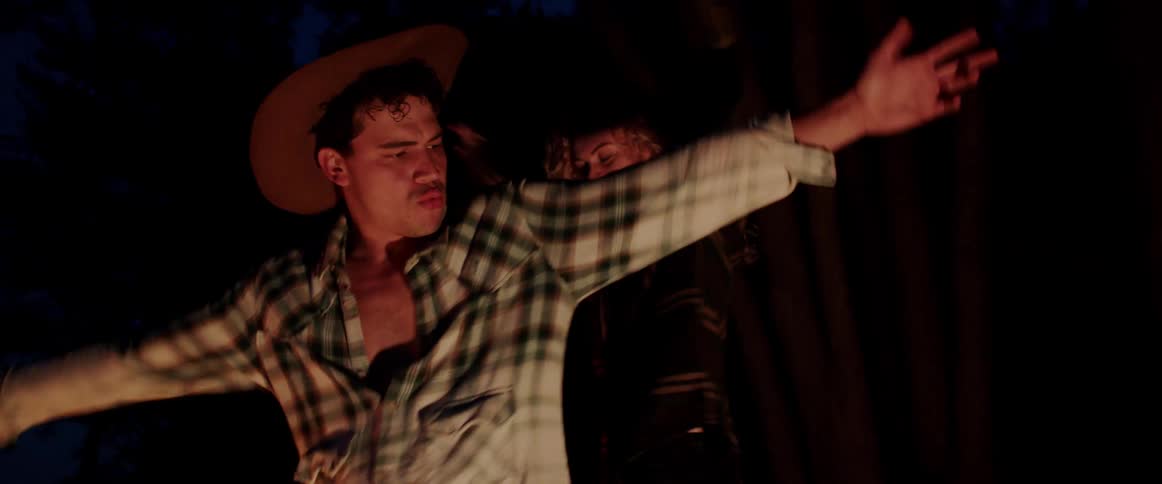 Man and woman dance outdoors in forest as firelight flickers on them