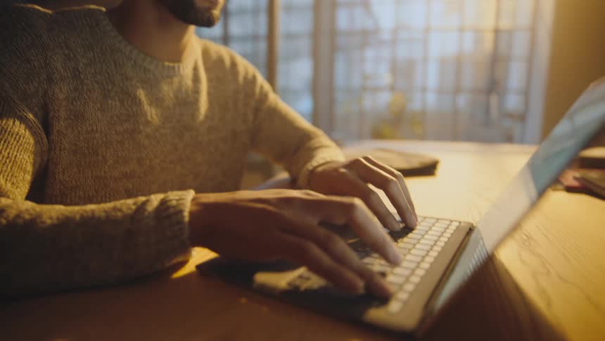 Man wearing a sweater and glasses types on a laptop while sitting at a table