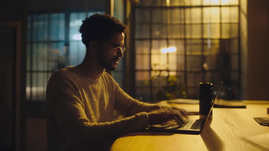 Time lapse of a  young man sitting at a desk working on a laptop at night