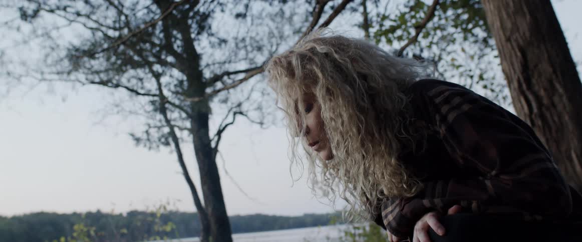Blonde woman crouches in front of a campfire next to a lake