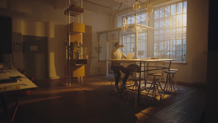 wide shot of a man sitting in an open office and looks at his computer and takes a drink