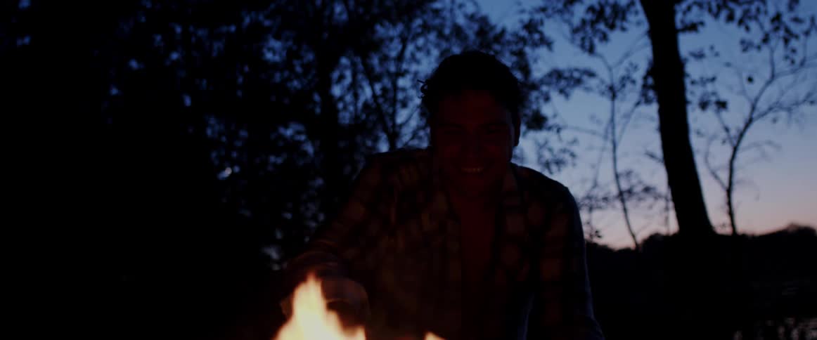 Man holds a cowboy hat as he crouches in front of a campfire at night next to the forest and lake