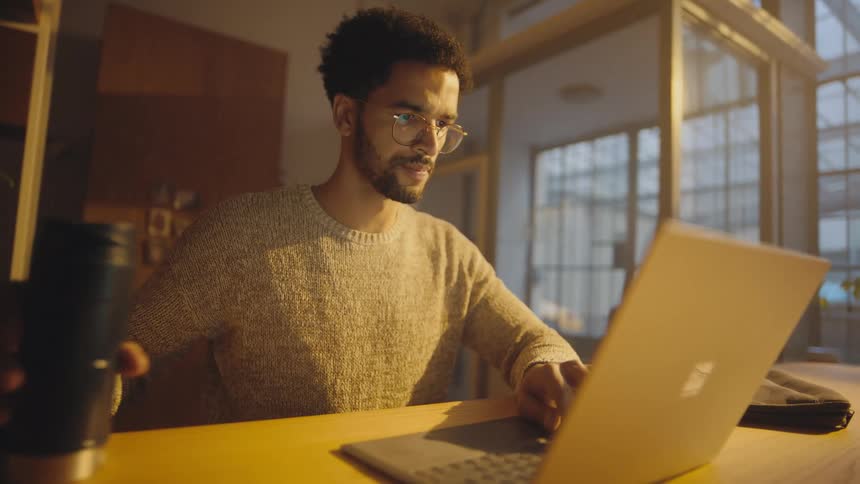 A man in an office types on his laptop and then picks up a cup and takes a drink