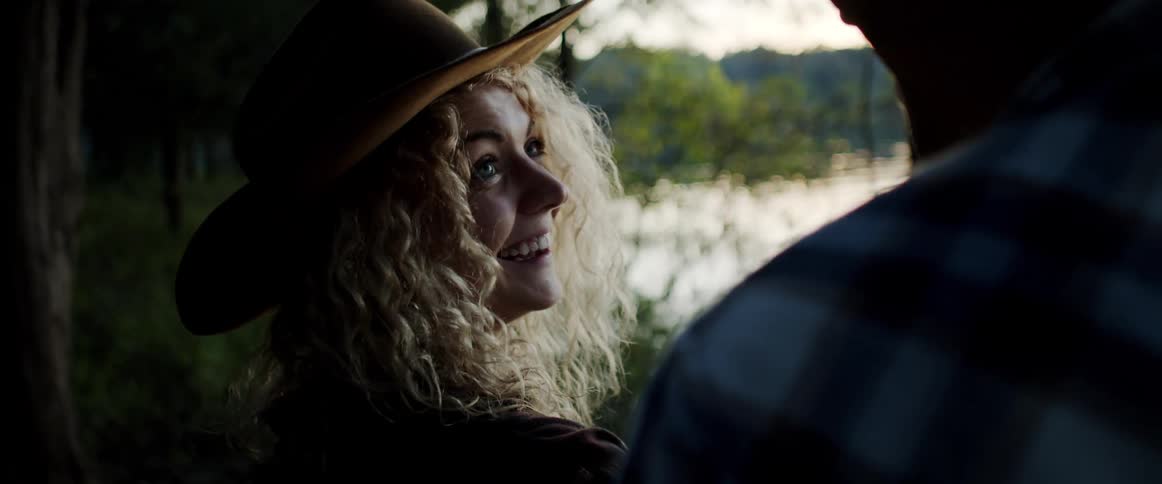 Young happy woman wearing a cowboy hat talking to her partner near a lake at morning