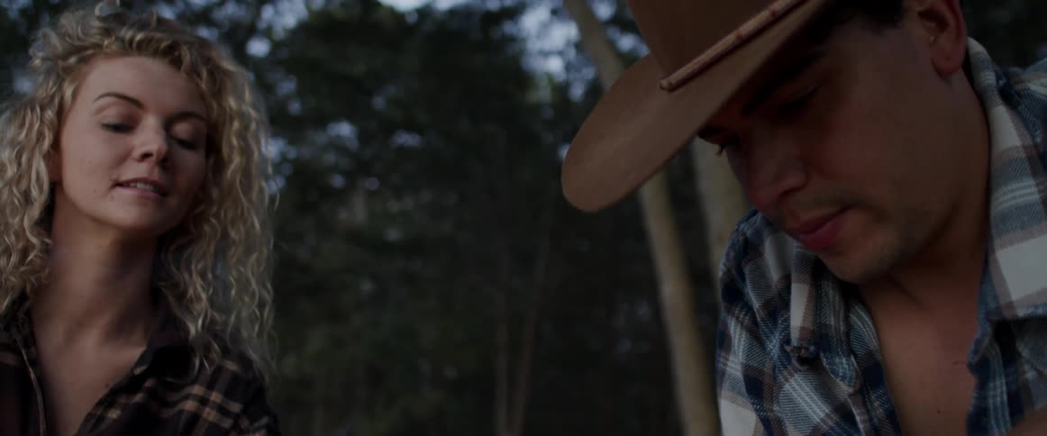 Blonde woman watches a man wearing a cowboy hat hold smoldering sticks and lower them while in the forest