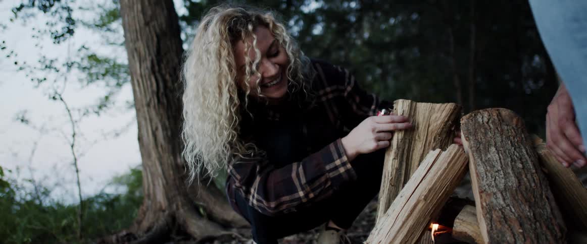 A woman and man stack firewood over a small campfire in the wilderness