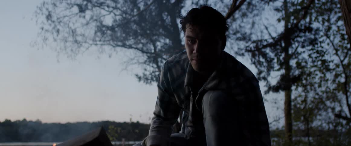 Man crouching in the forest ligths a campfire near a lake at evening
