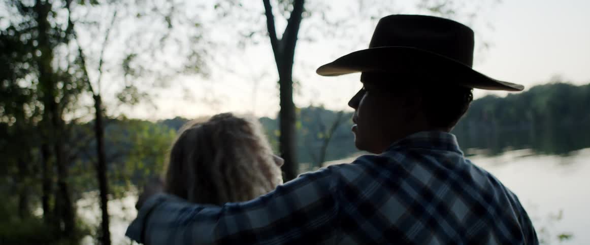 Man removes a cowboy hat from a womans head then puts it on his own head then hugs the woman as they both look out at a lake