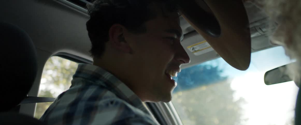 Blonde woman places a cowboy hat on a mans head while they travel in a car on a sunny day