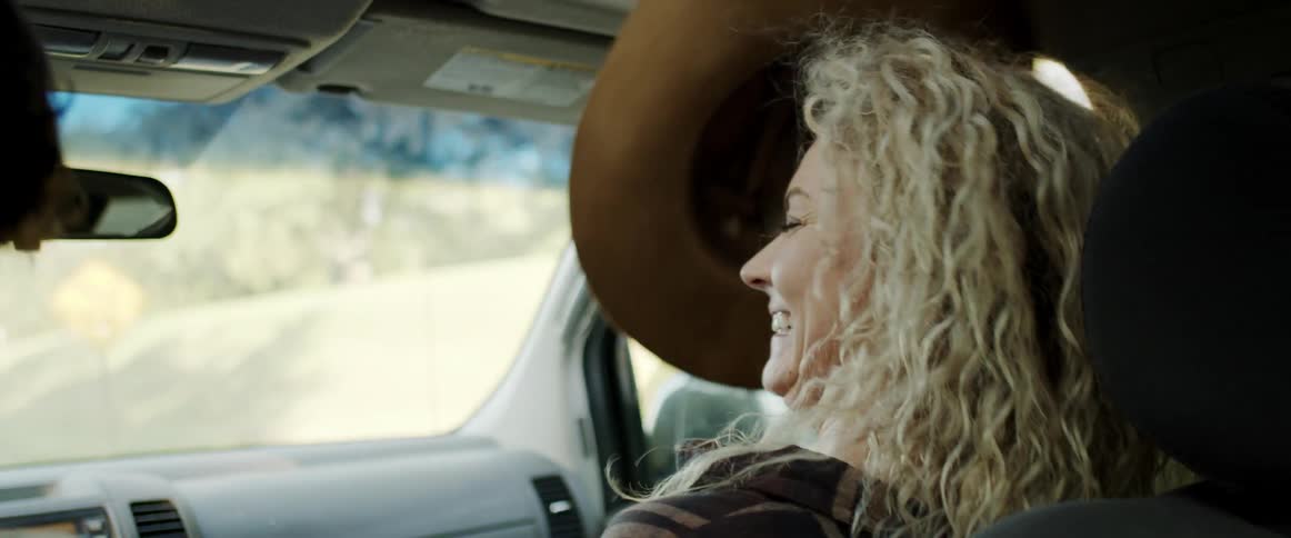 Blonde female passenger smiles and takes cowboy hat off male drivers head