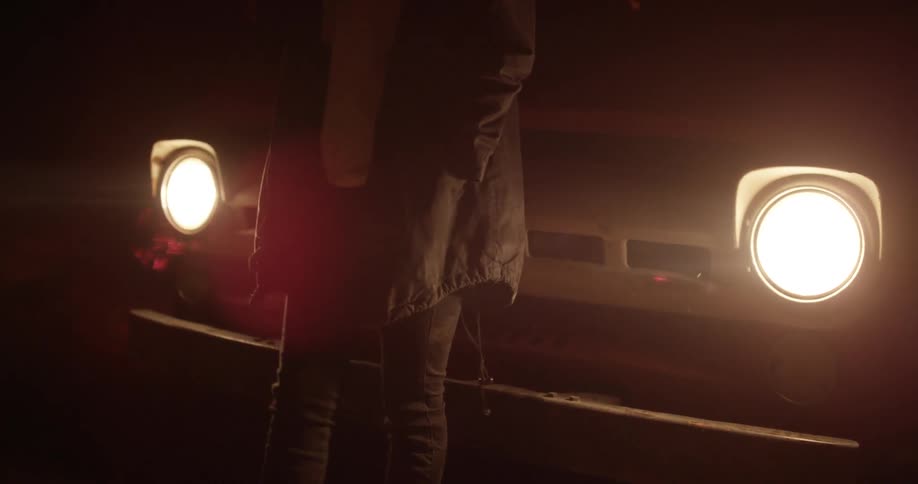 A person is standing in front of a vehicle as the headlights turn on