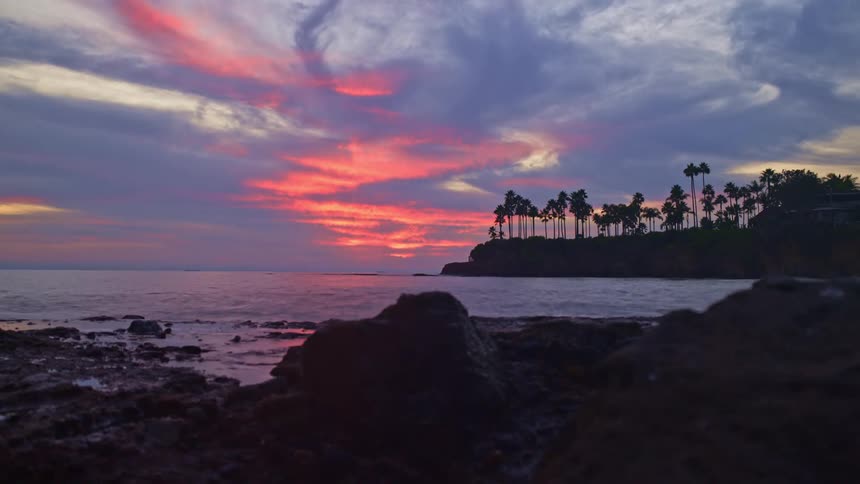 Rocks sit next to a lake as palm trees sit in the distance under a red sky at dusk and into the night