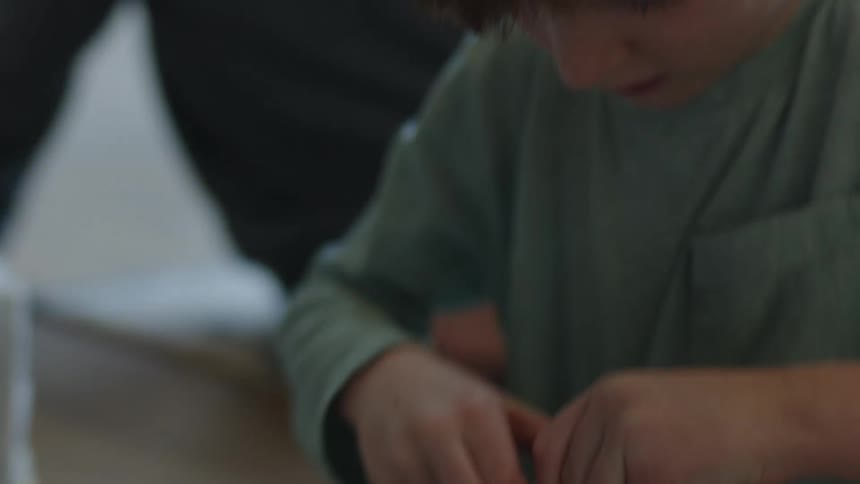Young boy trying to open a gift wrapped with a ribbon as an adult sits next to him and watches