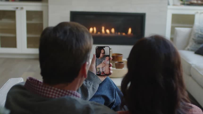 A man and woman sitting on the sofa talking to their daughter on a video call as she joyfully shows off her baby