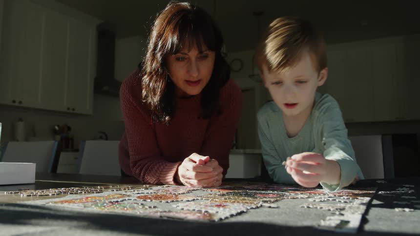 A mom leans on the table watching her young son put together a jigsaw puzzle