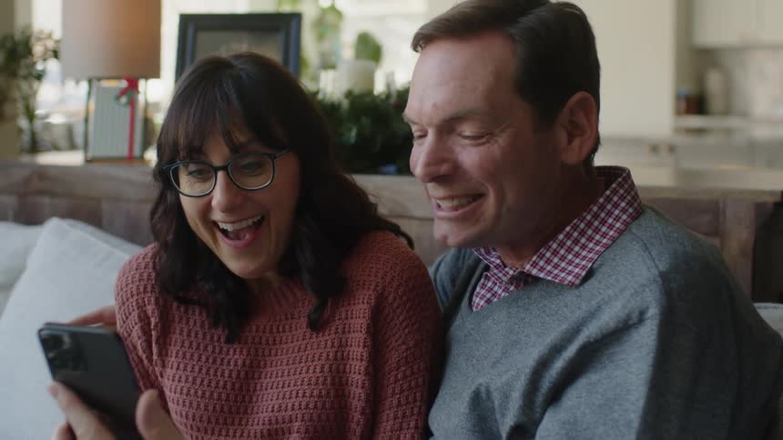 Happy couple sitting on a couch at home celebrate receiving good news in the phone