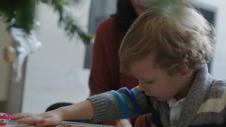 A little boy unwraps a gift under the christmas tree while mom sits beside him
