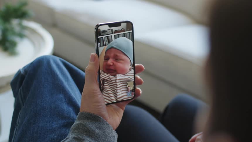 A man sitting with another person on a couch look at a crying baby on their smart phone