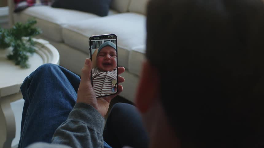 A man sitting in his living room holding a phone watching his baby grandson