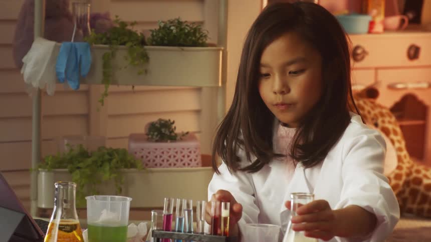 A young girl at home looks at a tablet as she pours liquid in to a beaker and out of a test tube during a science experiment