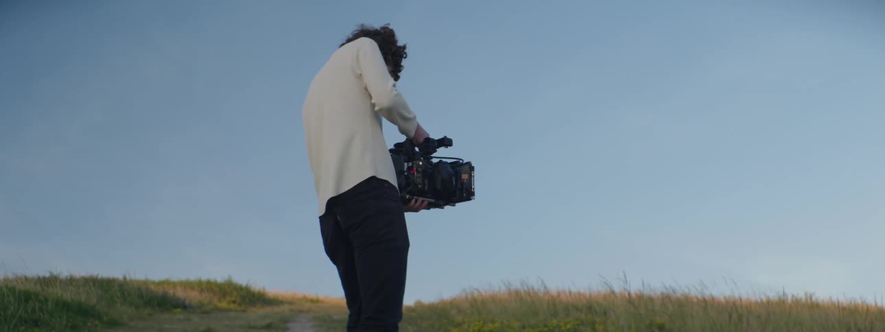 A man records video with a professional camera while standing in a field