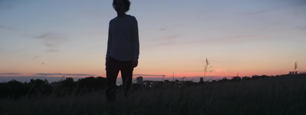 A man walks through a field outside a city at sunset