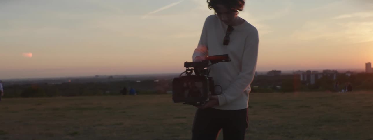 Cameraman with curly hair white sweater and black pants uses film camera in the field with city skyline in the background at sunset