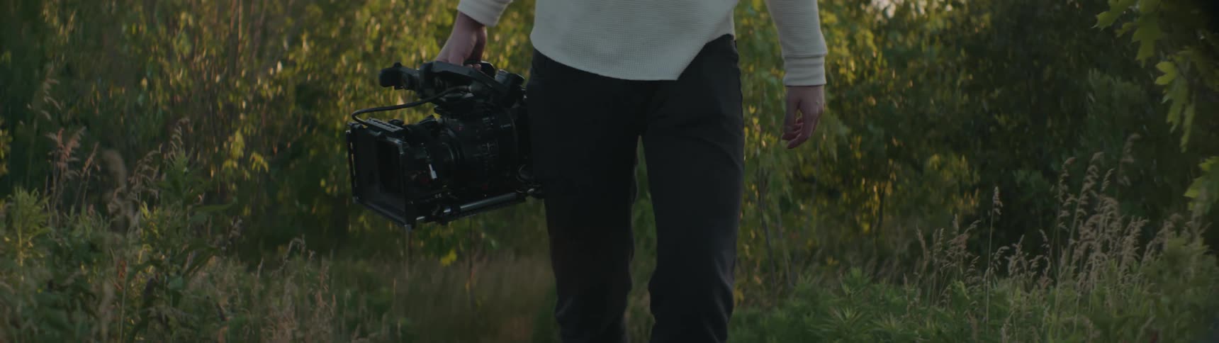 Cameraman with white sweater and black pants carries film camera in the forest