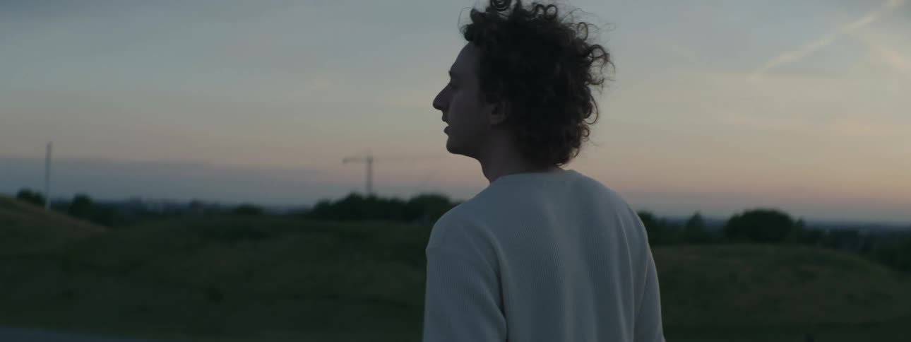 Cameraman with curly hair and white sweater walks downhill carrying film camera at sunset