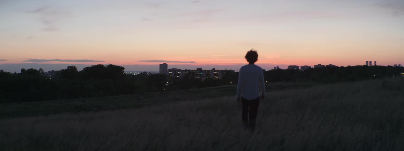 Man with curly hair white sweater and black pants walks downhill at sunset