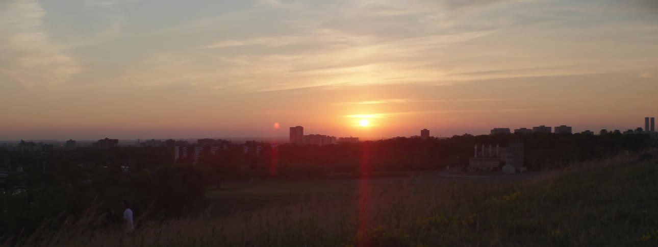 Man with white shirt stands on hill slope with city skyline in the background at sunset