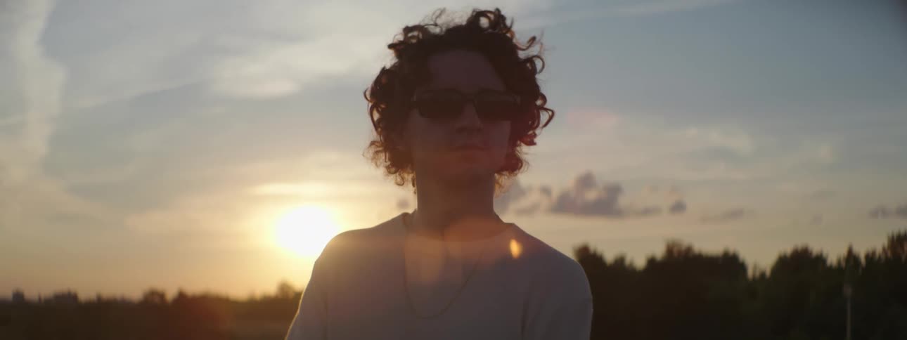 A man wearing sunglasses standing outside at sunset looking around