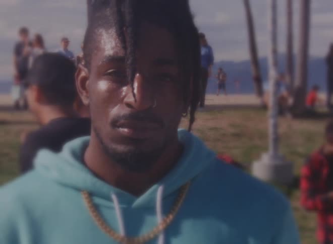 Man with dreadlocks nose piercing blue hoodie and necklace stares at camera at social event outdoors