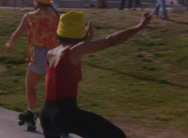 Two women with yellow wooly hat red shirt black leggings orange hat blouse and shorts roller skating at social event outdoors