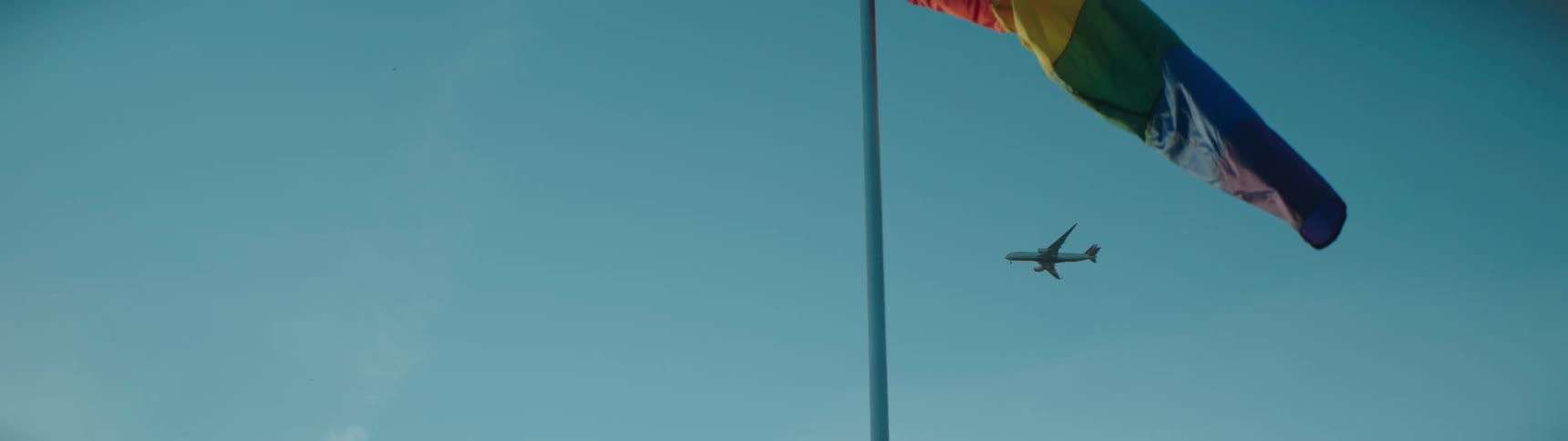 A rainbow windsock flutters in the wind as an airplane travels overhead