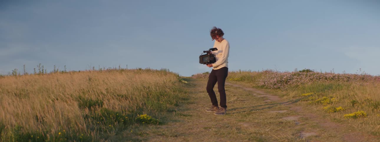 Filmmaker holds a camera while filming the horizon from a path in a field