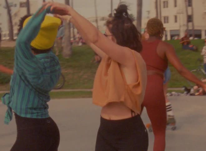 Two women dance together as a group of people roller skate in a park