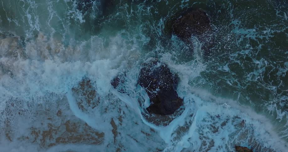 Waves crash against rock outcropping and nearby beach creating foam and sending spray into the air