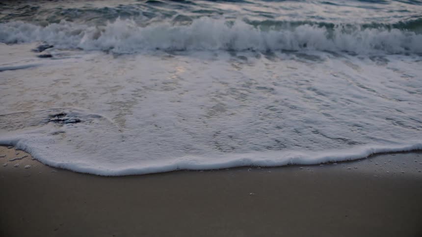 Ocean waves splash on a sandy shore with the wet sand reflecting the sky and clouds