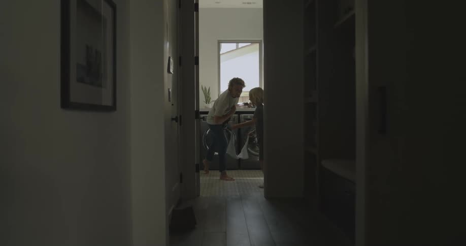A man playfully whips a boy with a towel causing the boy to run away down a corridor