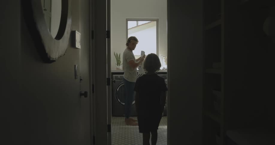 A man is putting towels in a washing machine and his son walks in the room and he laughs and hits his son with a towel