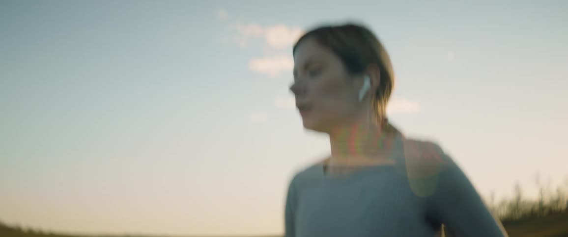 Woman wearing earbuds parallels sidewalk as she runs with forest on horizon