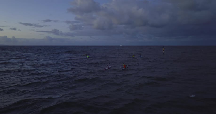 A group of kayakers row toward a marker with a flashing light at sunset