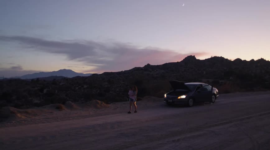 Mother walks away from car broken down on dirt road through scrub then turns to watch father walking around car